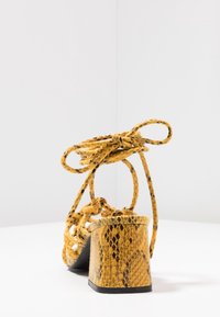 Żółte sandały na wysokim obcasie w print węża z grubym obcasem i plecionymi paskami wiązanymi w kostce. Teksturowana powierzchnia z błyszczącym wykończeniem.