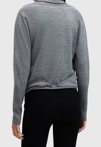 Szary sweter z długim rękawem o prążkowanej fakturze, z kołnierzem składanym i dwoma guzikami z tyłu. Modelka ma na sobie czarne spodnie.