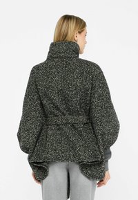Cappotto oversized a zig-zag bianconero con alto colletto e vita cintata. Texture morbida, maniche ampie e dettaglio plissettato sulla schiena.