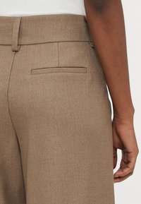 Brūnas piegrieztas bikses ar gludu struktūru, jostas cilpām un vienu aizmugurējo kabatu ar atveres dizainu, attēlots no aizmugures.