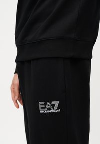 Άτομο που φοράει μαύρο παντελόνι φόρμας Emporio Armani EA7 και αντίστοιχο μαύρο φούτερ, δείχνοντας το πλευρικό λογότυπο στο παντελόνι φόρμας.