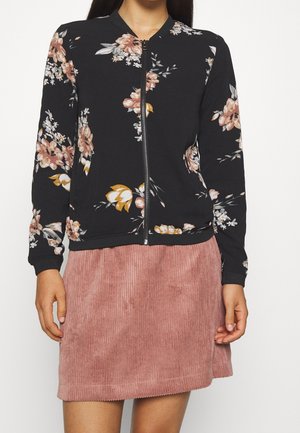Kvinde iført en sort blomsterprint lynlåstrøje og et støvet rosa kordnøjlskørt, vist fra halsen til midt på låret mod en hvid baggrund.