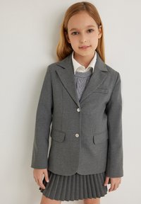Grijze blazer met een getailleerde pasvorm en twee voorzakken. Gepaard met een witte colbert met kraag en een grijze gebreide trui, gedragen met een geplooide rok.