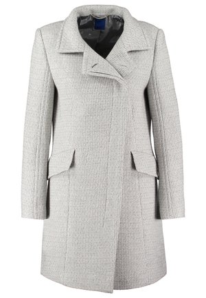 Femme portant un manteau gris clair texturé, une blouse en satin rose, un pantalon noir, des bottines noires, portant un sac à main noir à clous.
