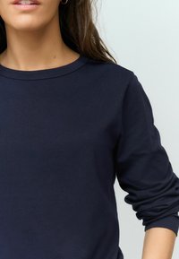 Tmavě modrá dlouhá košile s dlouhým rukávem, vyrobená z měkké látky, kulatý výstřih, s vyhrnutými rukávy a hladkou texturou. Minimalistický design, žádná viditelná loga.
