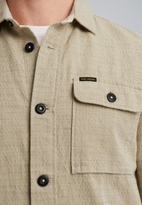 Beige, gestructureerd shirt met een knoopsluiting aan de voorkant, zwarte knopen en een borstzak met een zwart etiket. De kraag is plat en gestructureerd.