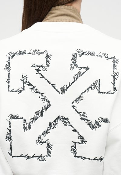 Personne portant un sweat-shirt blanc avec un texte en cursive noire formant un motif symétrique stylisé dans le dos.