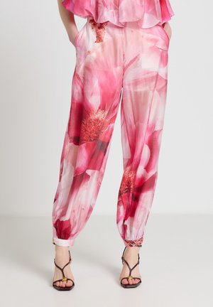 Femme portant un pantalon large rose à motifs floraux avec des bas élastiques et des sandales noires à talons hauts et brides, debout les mains dans les poches.