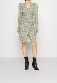 Grünes Blumen-Wickelkleid mit langen Ärmeln, V-Ausschnitt und seitlichem Schlitz, kombiniert mit schwarzen Stiefeletten mit klobigem Absatz und Schnalle.