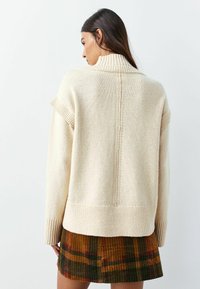 Beige strikket sweater med høj krave, ribbede manchetter og kant; har en centralt placeret sømdetalje. Kombineret med et flerfarvet ternet skørt.