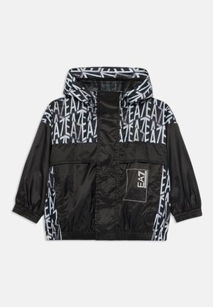 Černá větrovka s kapucí, zdobená geometrickým vzorem a šedým logem. Elastické manžety a přední kapsa s klopou.