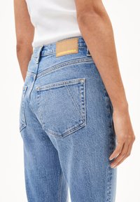 Jeans en denim bleu clair avec taille haute, deux poches arrière avec couture en diagonale, et un patch en cuir beige avec logo en relief sur la ceinture.