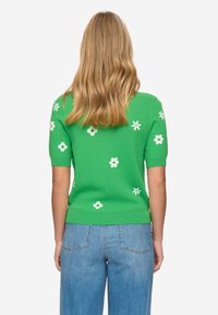 Zelený svetr s krátkým rukávem z pletené látky, zdobený bílými květinovými motivy a vroubkovaným lemem. Nošený s modrými džínami.