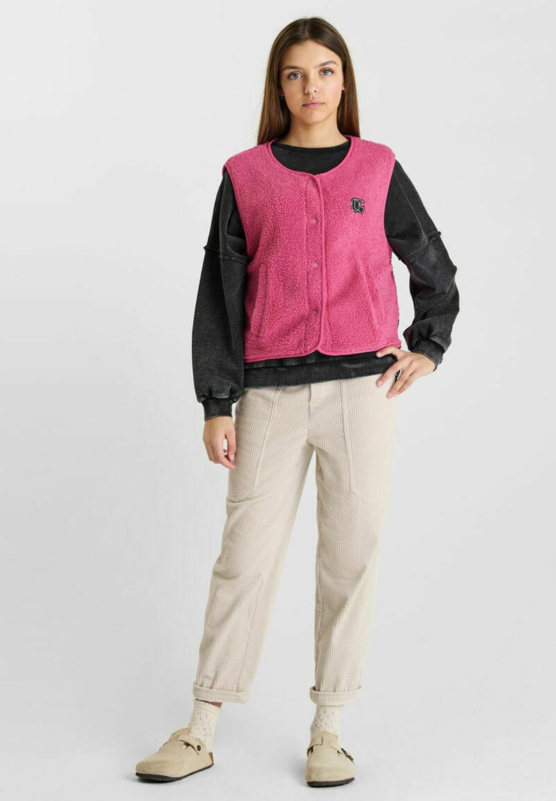 Fleece vest in felroze met een ronde halslijn, knopen aan de voorkant en zijzakken. Draagt over een zwart langemouwen shirt en beige corduroy broek.