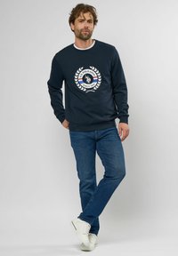 Marineblaue Sweatshirt mit einem runden Logo-Design in Weiß, Blau und Rot, kombiniert mit blauen Jeans und weißen Sneakers. Glatter Stoff, lässige Passform.