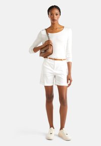 Bílý dlouhý rukáv, upravené bílé šortky, oranžový pásek, hnědá crossbody taška s řetězem a krémové tenisky. Hladké textury po celém outfitu.