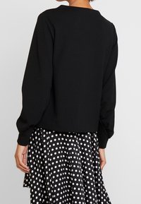 Musta lyhyt neulepusero, jossa pitkät hihat, yhdistetty mustavalkoiseen polkadotpaitahameeseen. Kankaan rakenne on teksturoitu ja kaula-aukko pyöreä ja yksinkertainen.