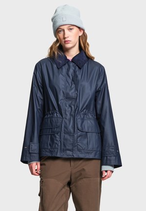 Jeune femme portant une veste bleu marine avec de grandes poches et un bonnet tricoté bleu clair, debout devant un fond uni.