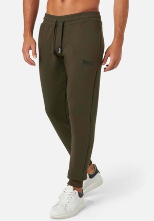 Olivalgrüne Sweatpants mit elastischem Bund und Kordelzug, versehen mit einem Seitenstreifen und einem Logo auf dem Oberschenkel. Kombiniert mit weißen Sneakers.