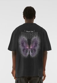 Tricou oversize negru, cu un grafic mare și detaliat al unei fluturi mov pe spate și text alb "migonzares" deasupra.