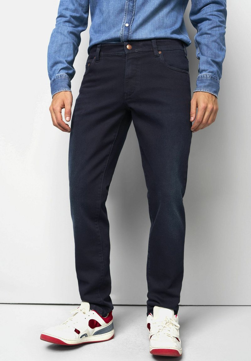 Dunkelblaue Jeans mit enger Passform, ausgestattet mit einem klassischen Fünf-Taschen-Design. Getragen mit einem hellblauen Denim-Hemd und weißen Turnschuhen.