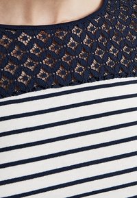 Collo in pizzo blu navy con motivo geometrico sopra un tessuto a strisce bianche e blu navy. Texture morbida, vestibilità casual e design con colletto tondo.