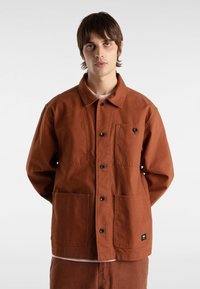 Bruine canvas jas met een knoopsluiting, twee voorzakken en een klein logotag. Gladde textuur met een ontspannen pasvorm.
