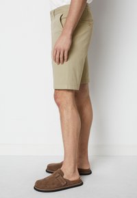 Beige cotton shorts with side pockets, sitting above the knee. Worn with brown suede slip-on shoes featuring a strap and textured sole.