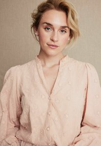 Ljust rosa blus tillverkad av texturerad tyg, med blommig broderi, en volangkrage och knappar fram. Långa puffärmar.