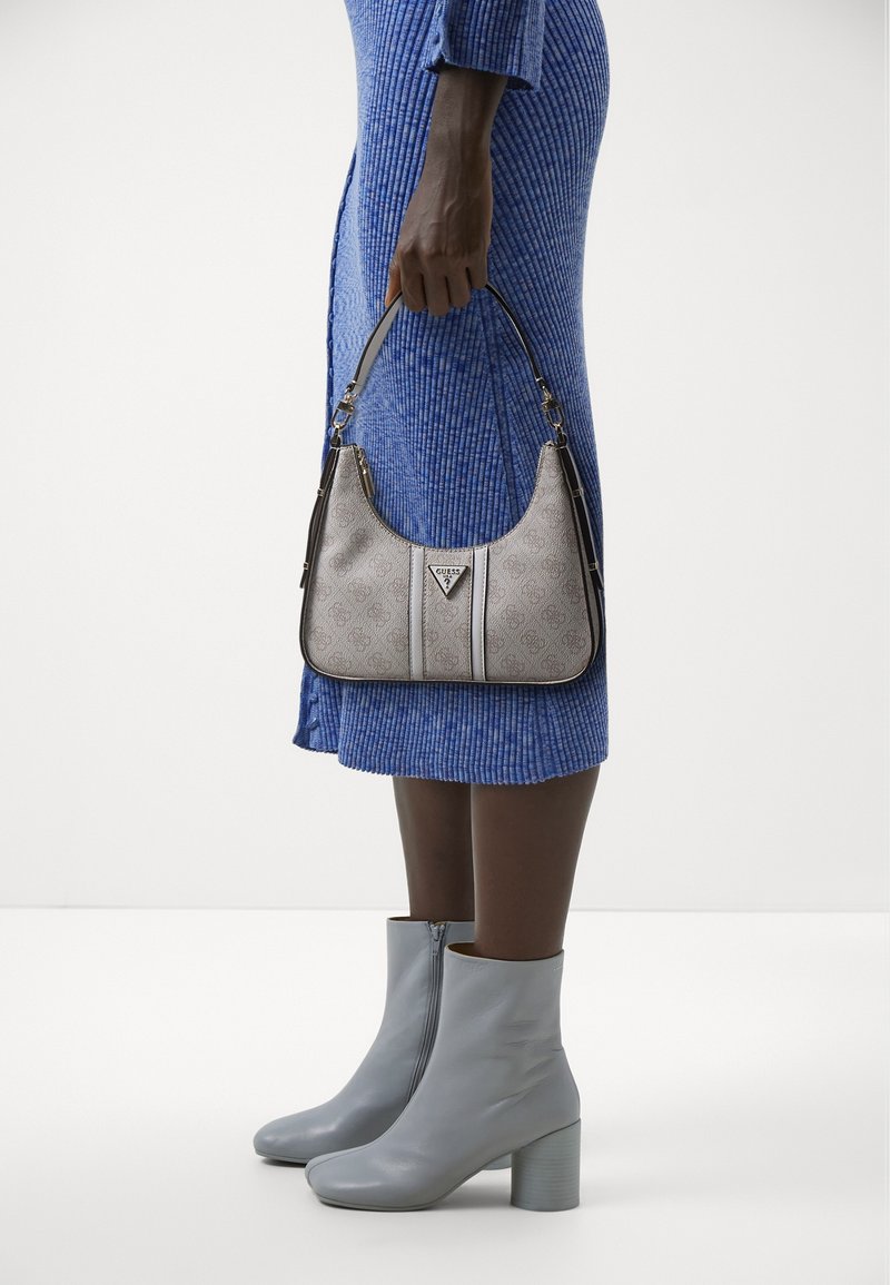Femme en robe côtelée bleue tenant un sac à main beige à motifs, portant des bottines bleu clair à talons blocs, debout sur un fond blanc.