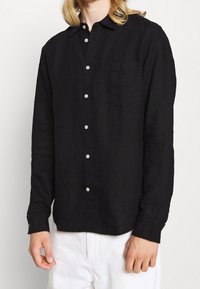 Černá dlouhá rukávová košile vyrobená z lehkého materiálu, s knoflíkovým zapínáním vpředu, jednou náprsní kapsou a knoflíkovými manžetami.