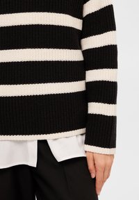 Il maglione lavorato a maglia presenta righe orizzontali bianche e nere, una texture a coste e una vestibilità rilassata con una camicia bianca a strati sotto.
