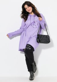 Lavendel lange hoodie met trekkoord taille, gecombineerd met zwarte broek met witte tekst, en zwarte studded enkellaarsjes. Zwarte gewatteerde handtas.
