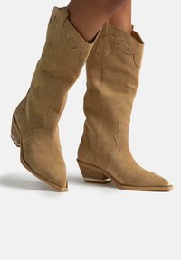 Stivali cowboy in suede beige, alti fino al ginocchio, con punta affusolata, design scallop e tacco basso impilato, dotati di suola testurizzata.