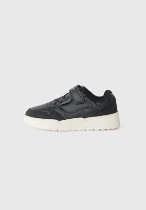 Sort lavtop sneaker med hvid sål, velcrorem og snøredetaljer vist fra siden på en ensfarvet lysegrå baggrund.