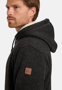 Felpa con cappuccio in tessuto grigio scuro con un aspetto texture, caratterizzata da un morbido rivestimento bianco e un'etichetta in pelle sulla spalla.