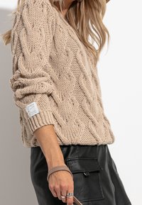 Beige stickad tröja med kabelmönster, ribbad nederdel och uppviktärmar; matchad med en svart läderkjol och handledsaccessoar.