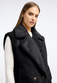 Czarna kamizelka z futerka syntetycznego z szerokim kołnierzem, podwójnymi guzikami, bez rękawów oraz miękką teksturą na jasnym tle.