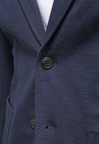 Närbild av en marinblå kavaj med svarta knappar fäst över en ljusgrå skjorta, som visar tygtextur och sömnadsdetaljer.