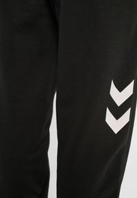 Zwarte sportbroek van zachte stof, met een wit chevronpatroon op het onderbeen. Gladde textuur, ontspannen pasvorm.