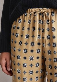 Pantalon beige à motif avec une taille à cordon, présentant un design géométrique en bleu et noir, fabriqué en tissu lisse avec une coupe décontractée.