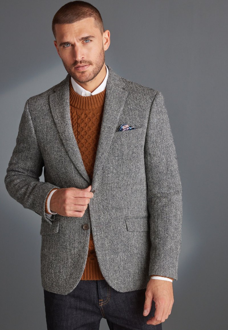 Grijze visgraatblazer met ingesneden revers, eronder een gebreide bruine trui met structuur, en een patroon zakdoek. Slim-fit ontwerp, knoopsluiting.