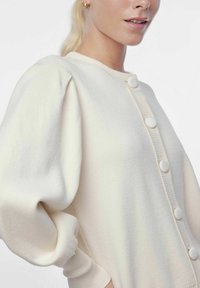 Crèmekleurige cardigan met een ronde hals, geribbelde afwerking en grote met stof beklede knopen; heeft pofmouwen en een zachte textuur.