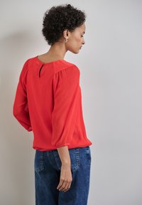 Rote, strukturierte Bluse mit Dreiviertel-Puffärmeln und einem Ausschnitt auf der Rückseite, kombiniert mit blauen Denim-Jeans.