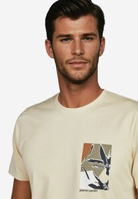 Beige Baumwoll-T-Shirt mit einem rechteckigen floralen Druck in Schwarz, Weiß und Erdtönen auf der linken Brustseite, mit rundem Halsausschnitt.