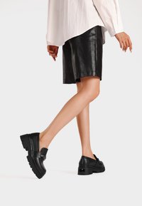 Scarpe in pelle nera con suola spessa, design slip-on e fascia decorativa. Indossate con una camicia bianca e pantaloni corti neri per creare contrasto.