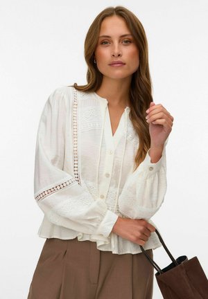 Mujer con largo cabello castaño que lleva una blusa blanca de encaje y pantalones marrones, sosteniendo un bolso marrón oscuro sobre un fondo blanco.
