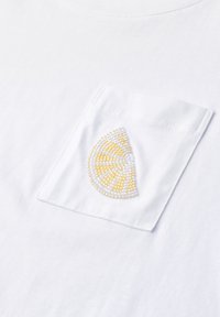 Tissu blanc avec une petite poche cousue présentant un motif perlé d'une tranche de citron jaune à l'avant.