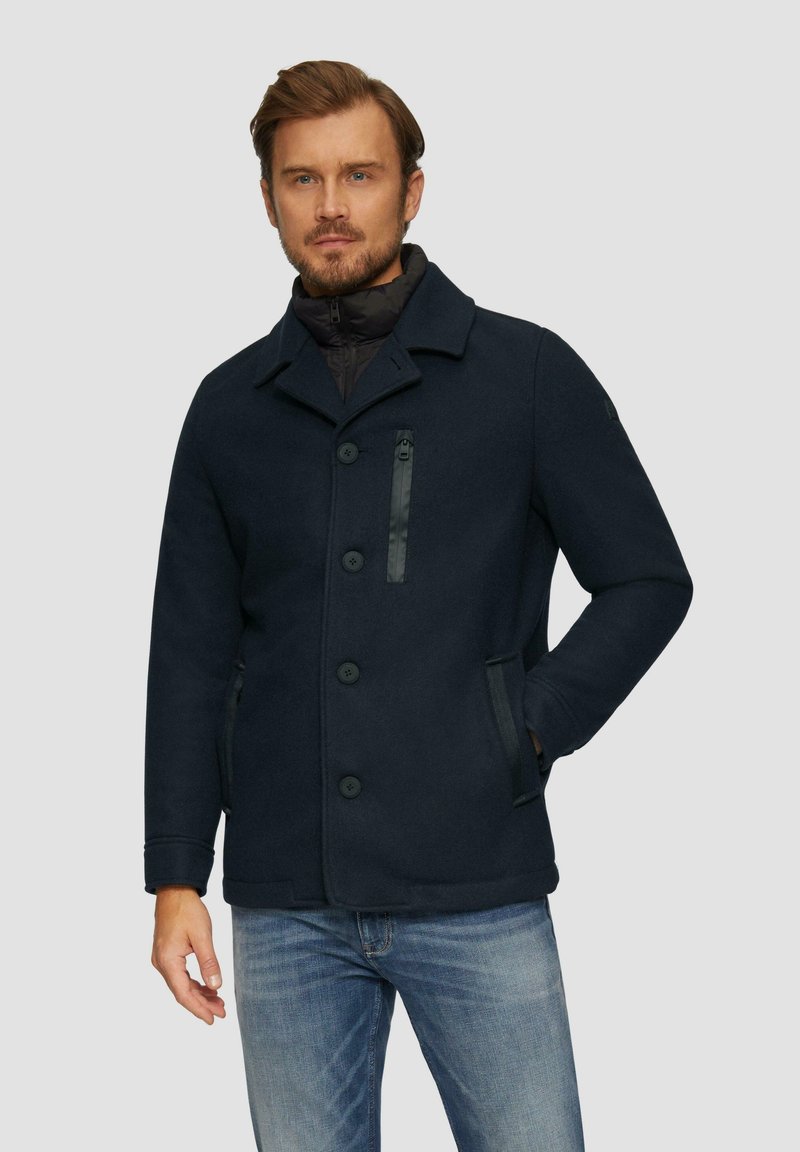 Dunkelblauer Wollmischgewebe-Mantel mit einem Reverskragen, vorderen Knöpfen, einer Reißverschlusstasche auf der Brust und Seitentaschen. Getragen über einem schwarzen gepolsterten Innenfutter.