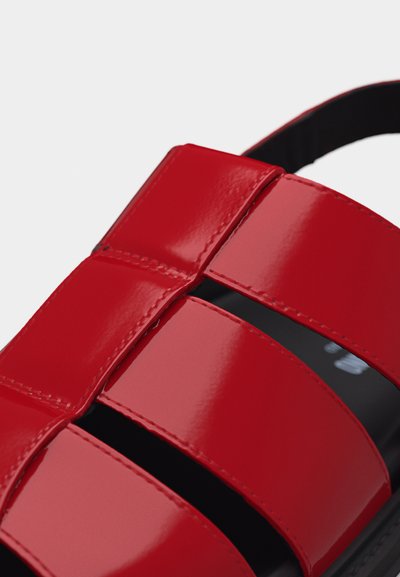 Κοντινή λήψη λαμπερής κόκκινης δερμάτινης σαγιονάρας με φαρδιές λουριά και εμφανές μαύρο στρίφωμα από κάτω.